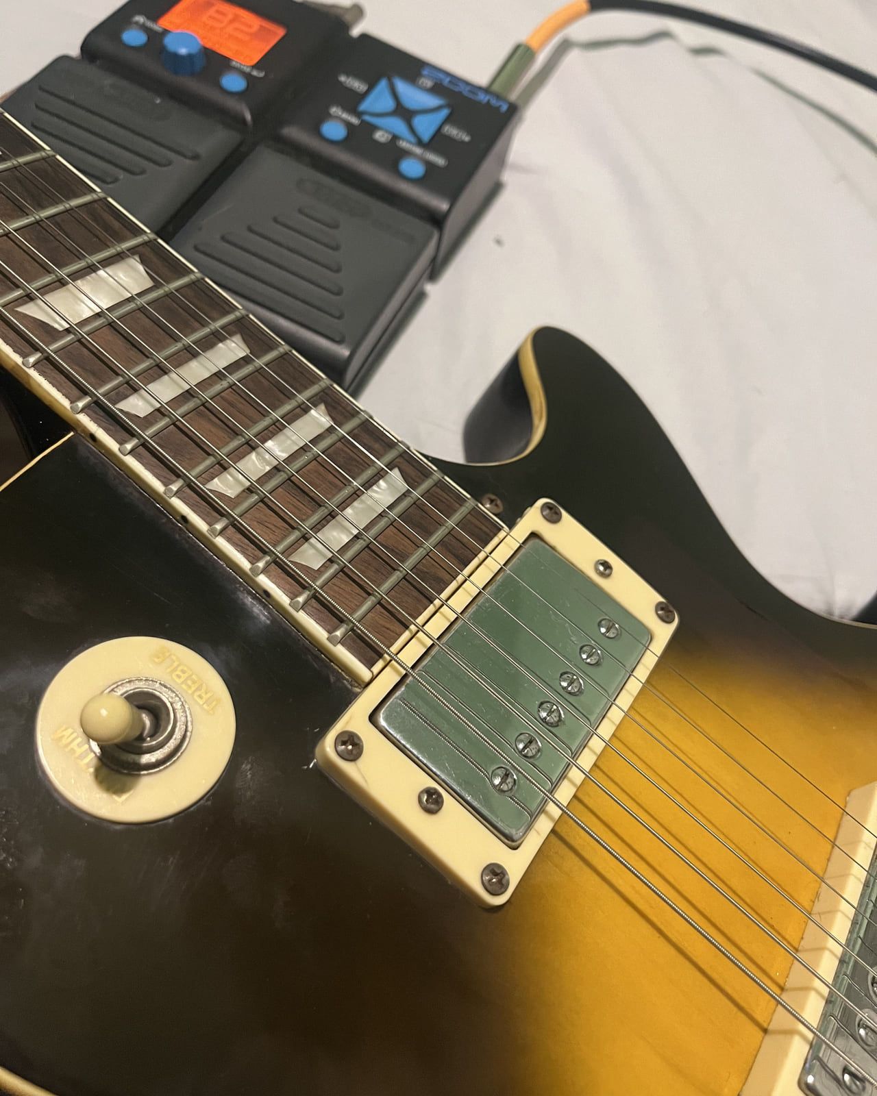Close de uma guitarra Les Paul sunburst com captador de capa cromada e frisos creme, apoiada ao lado de dois pedais Zoom de LEDs azuis e visor laranja