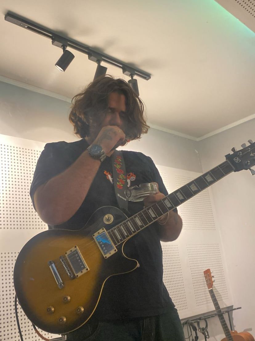 TazTheDoctor em pé num estúdio de paredes claras, tocando uma guitarra Les Paul sunburst, com o cabelo comprido caindo no rosto e um violão encostado ao fundo