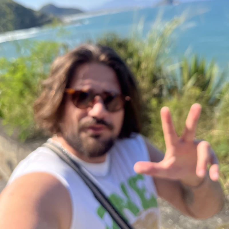 Selfie desfocada de TazTheDoctor de óculos escuros num mirante, com o mar e morros ao fundo
