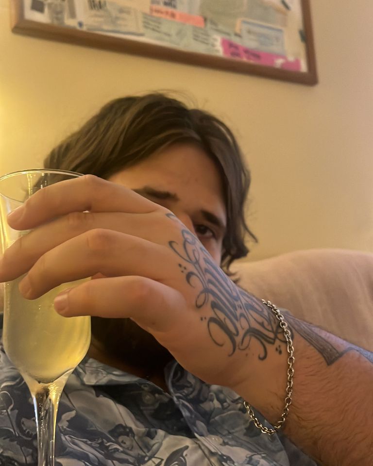 TazTheDoctor sentado, de camisa estampada, erguendo uma taça; a mão tatuada cobre parte do rosto